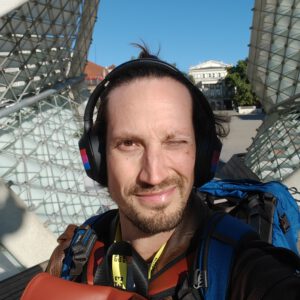 Selfie of Martin Tilo Schmitz. A white guy with trimmed beard and long brown hair in a bun, squinting against the sun, wearing big over-ear noise canceling headphones with the bi-flag taped on them.