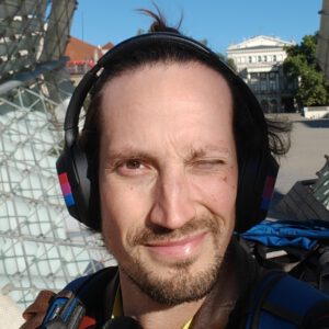 Selfie of Martin Tilo Schmitz. A white guy with trimmed beard and long brown hair in a bun, squinting against the sun, wearing big over-ear noise canceling headphones with the bi-flag taped on them.